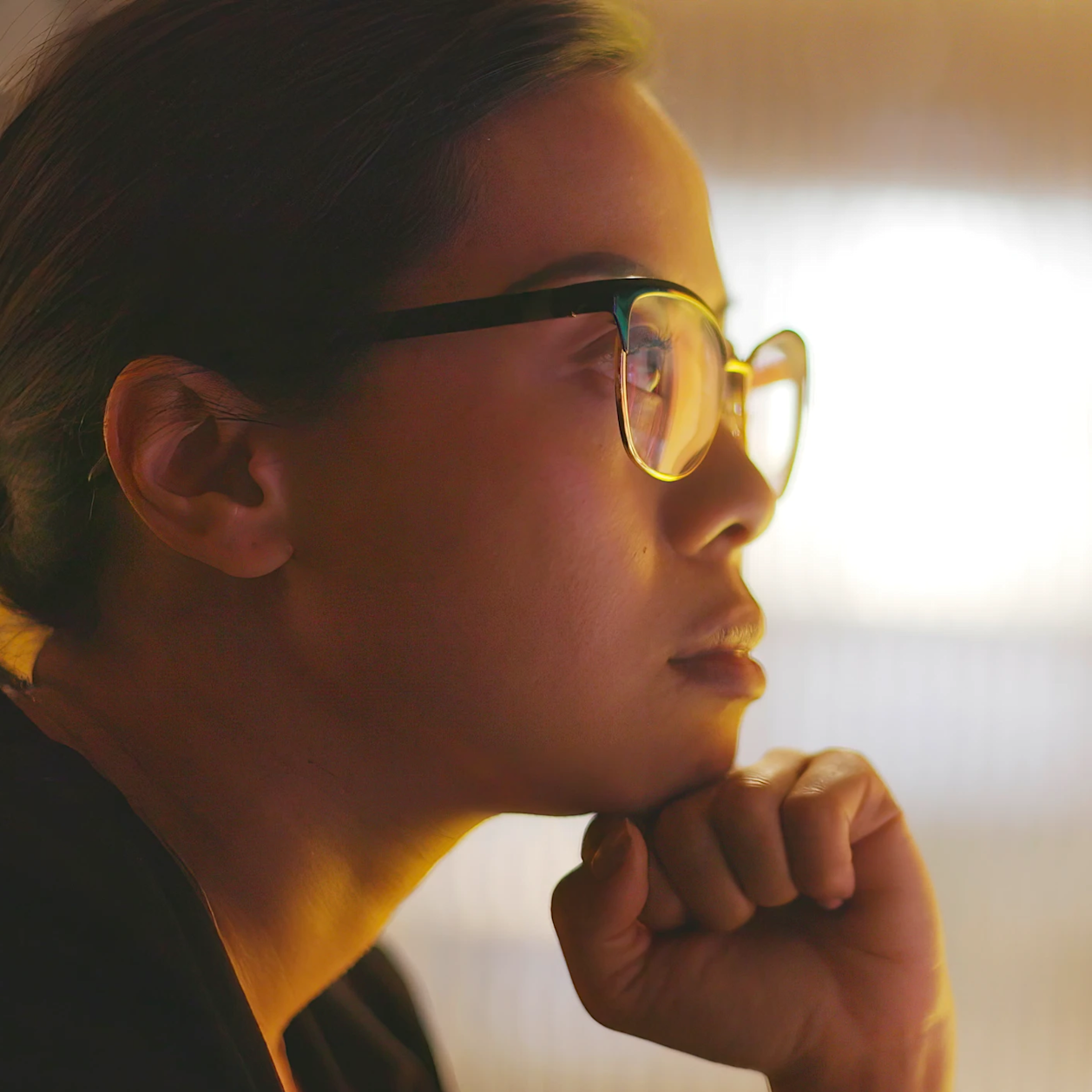 Close-up side profile of a person wearing glasses, resting their chin on their fist, looking thoughtfully into the light source.