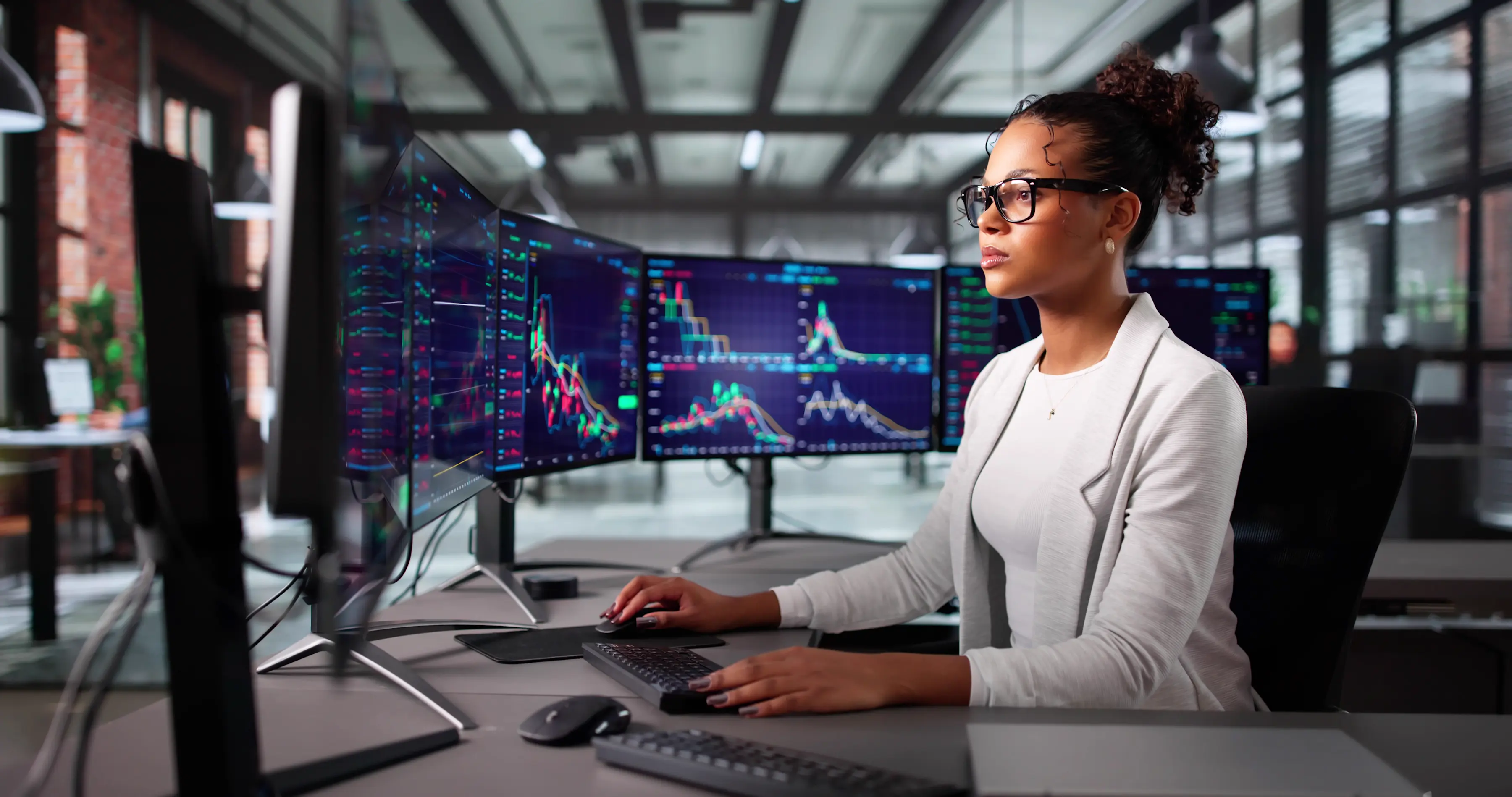 A woman wearing glasses seated at a desk working at a keyboard, with multiple monitors displaying colourful financial charts and data.