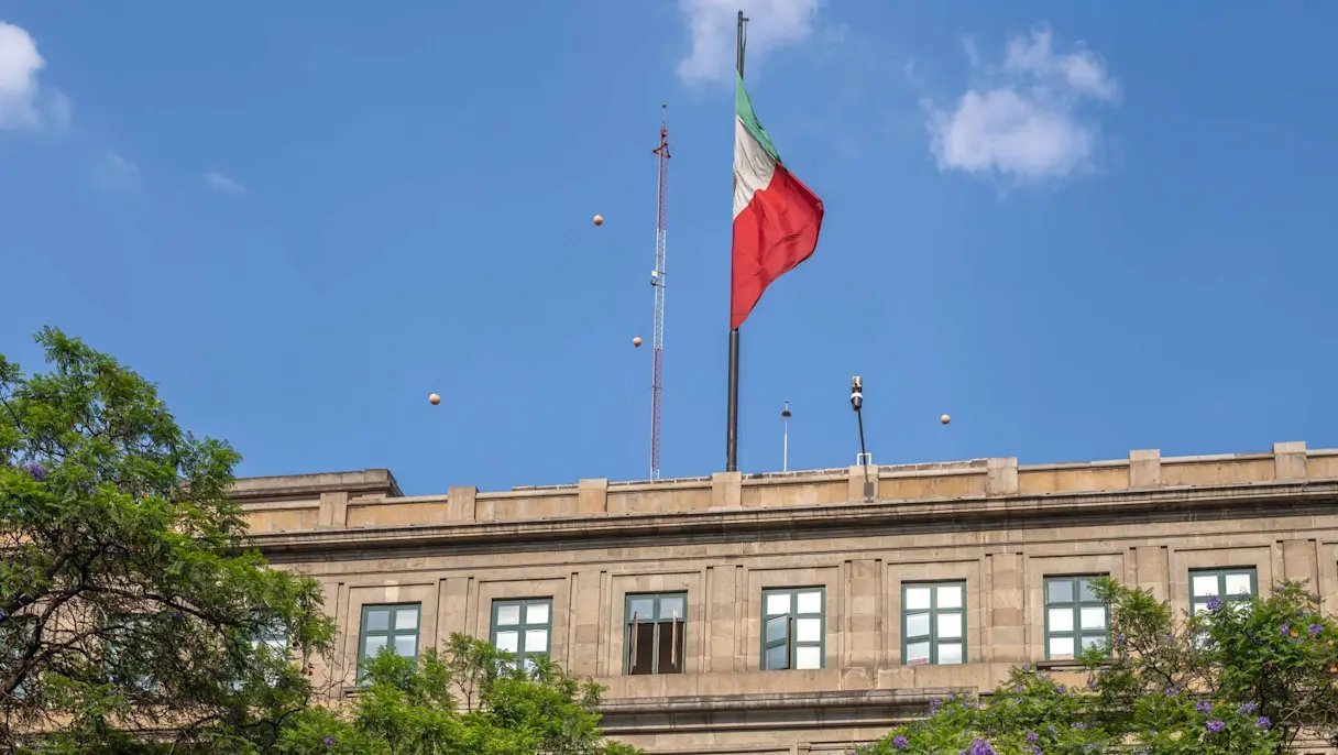 Mexican flag flying above a stone government building with green-framed windows, framed by trees under a blue sky.