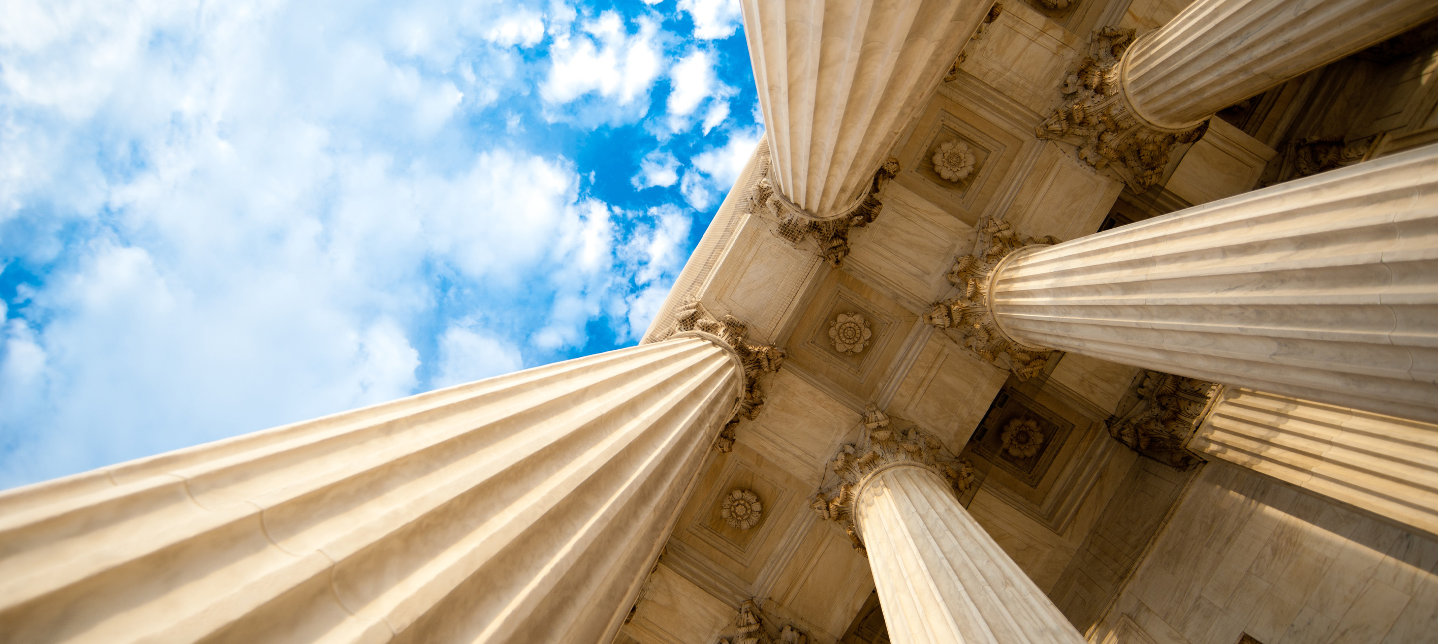 Low-angle view of towering classical stone columns and ornate architectural details against a bright blue sky with scattered clouds.