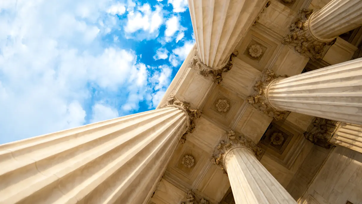 Low-angle view of towering classical stone columns and ornate architectural details against a bright blue sky with scattered clouds.