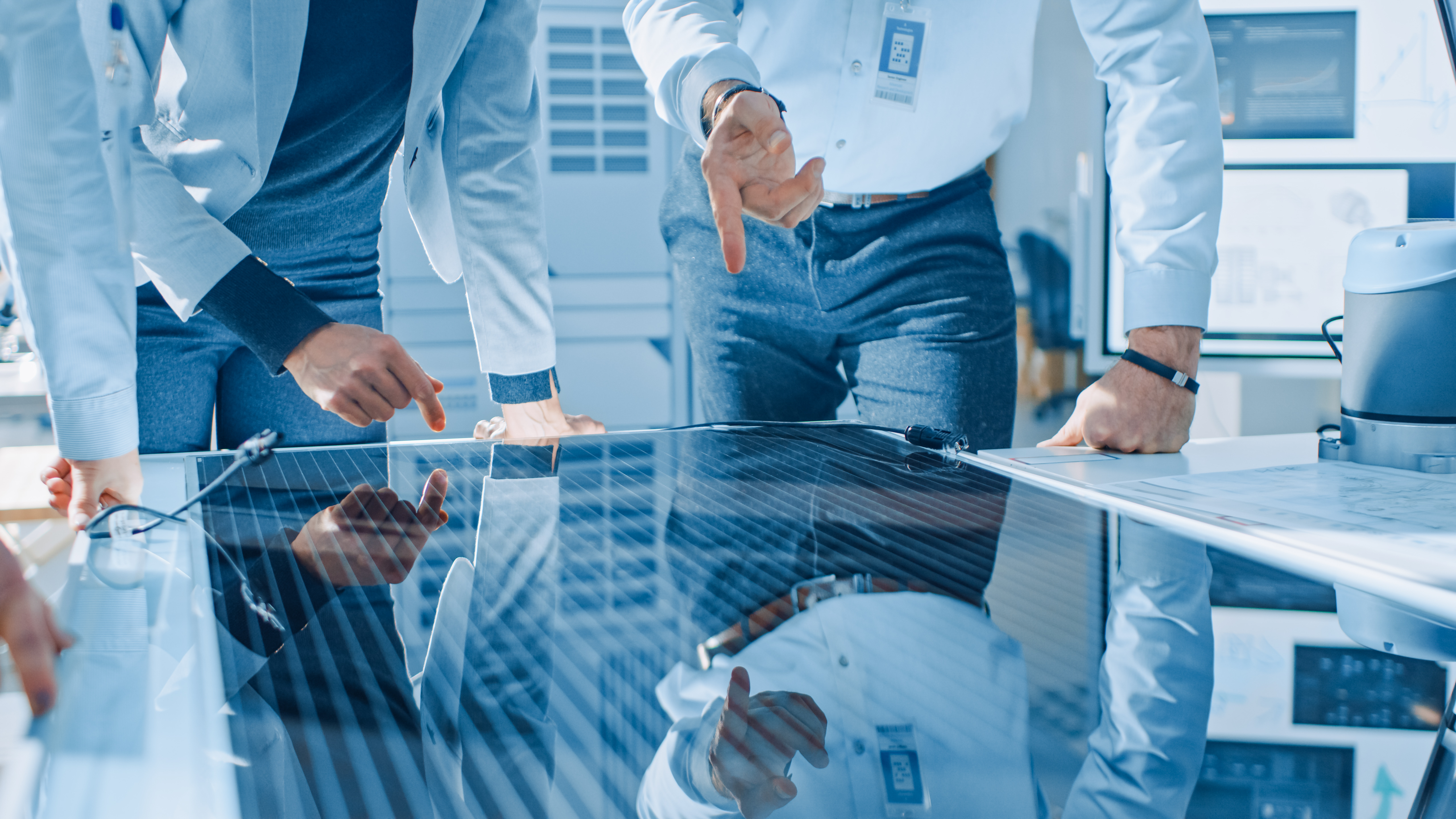 Two professionals in light blue shirts pointing at a large reflective solar panel on a table in a bright office setting.