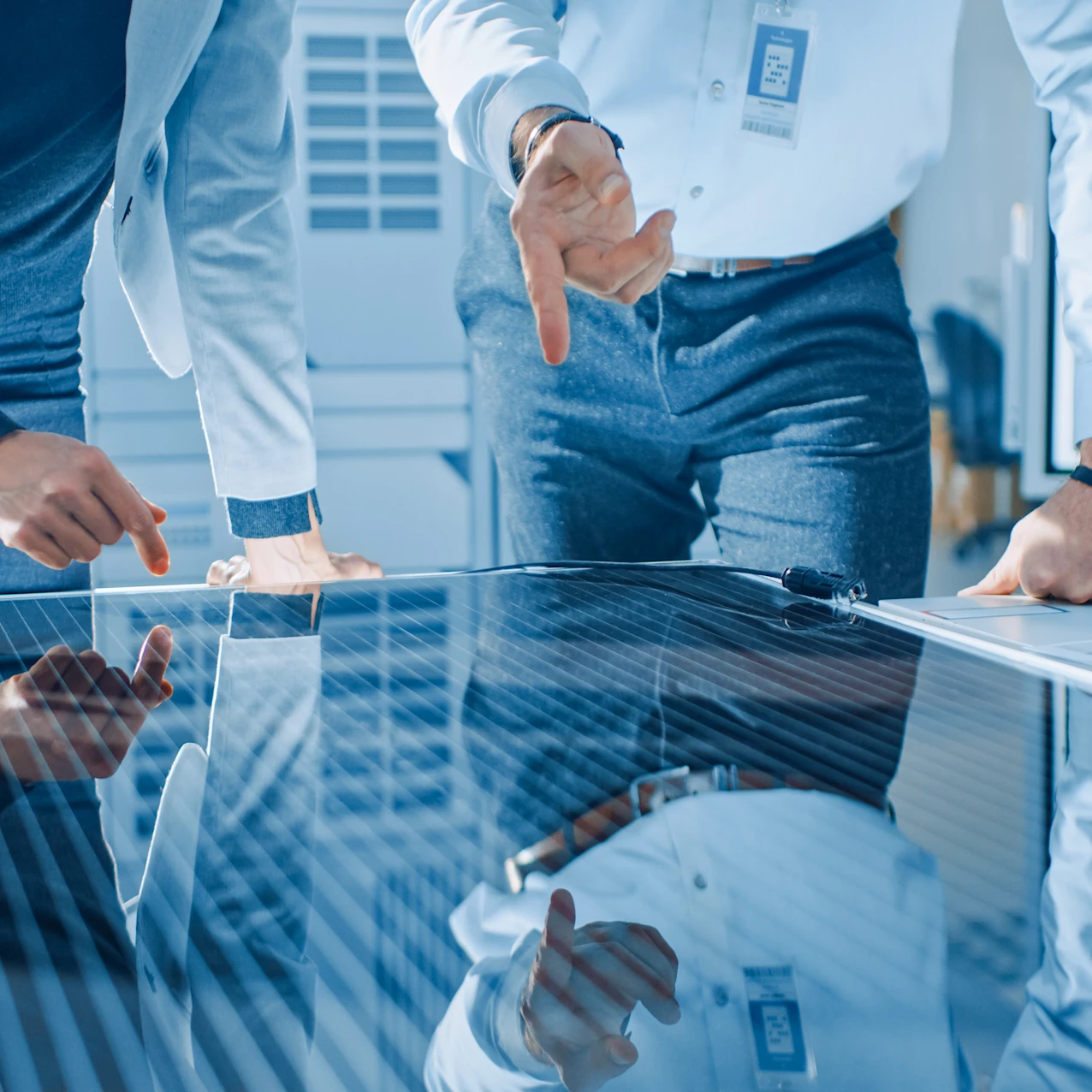 Two professionals in light blue shirts pointing at a large reflective solar panel on a table in a bright office setting.