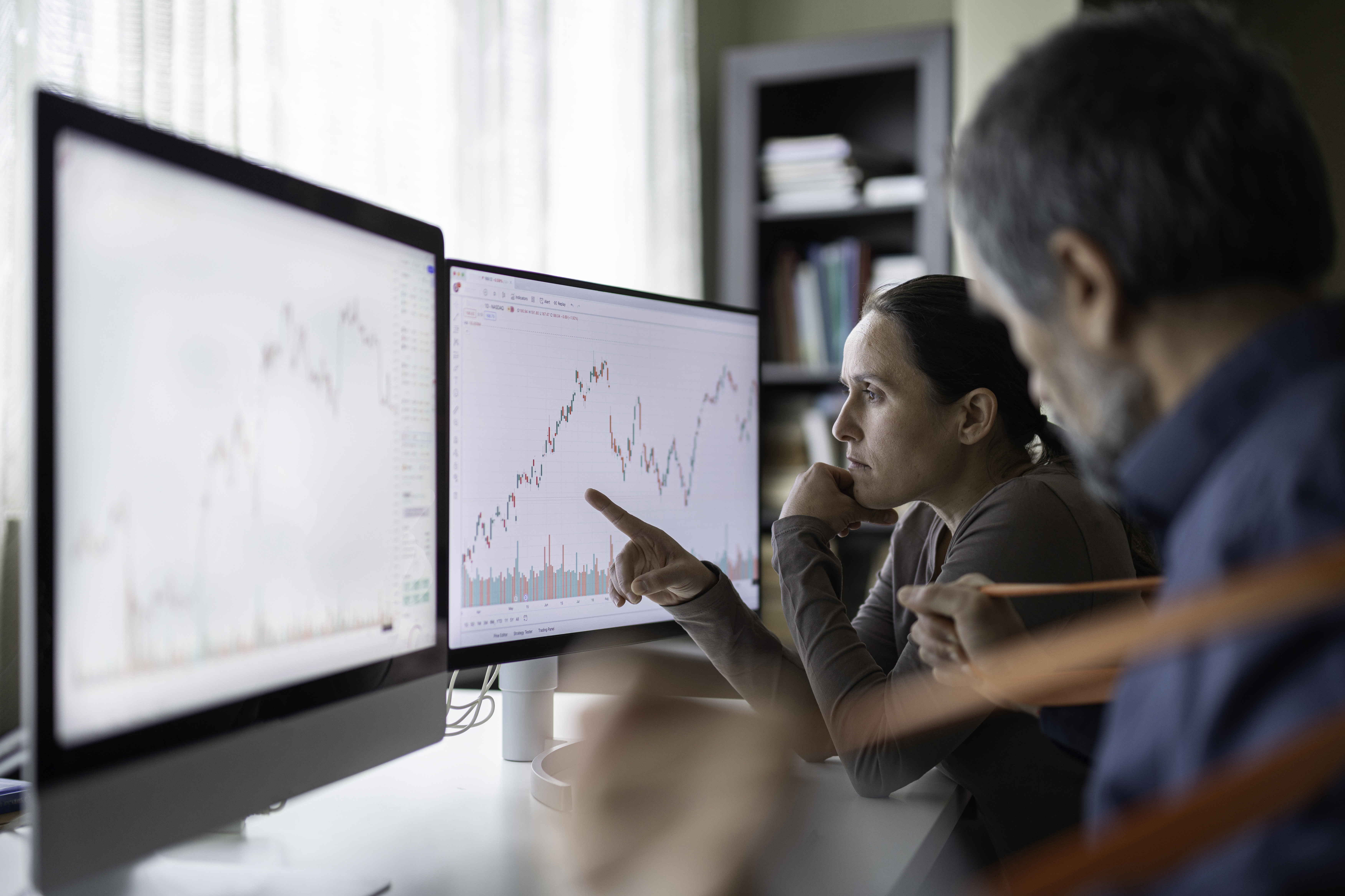 Two people analyzing stock market charts on dual computer monitors in a modern office setting.