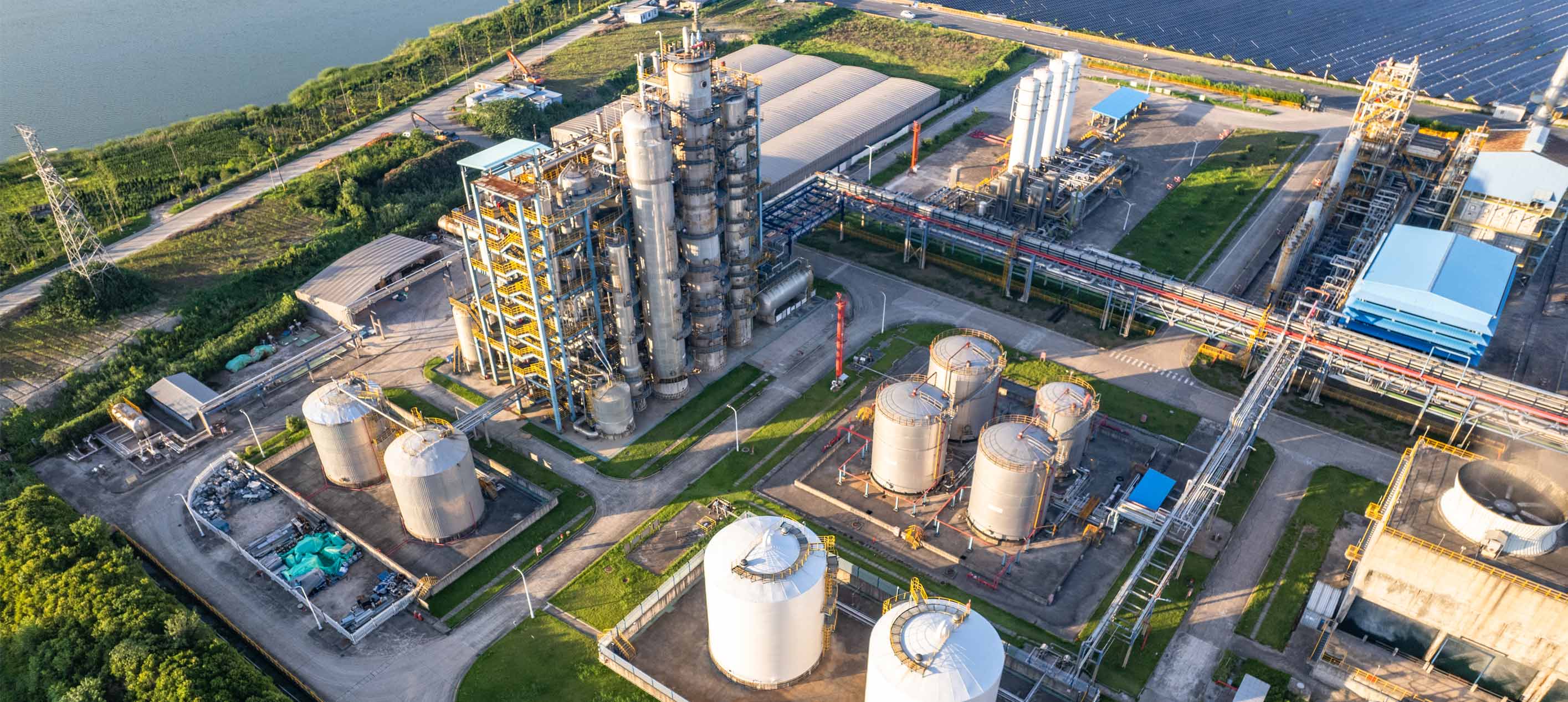 Aerial view of an industrial plant with tall processing towers, white storage tanks and pipelines beside a body of water.
