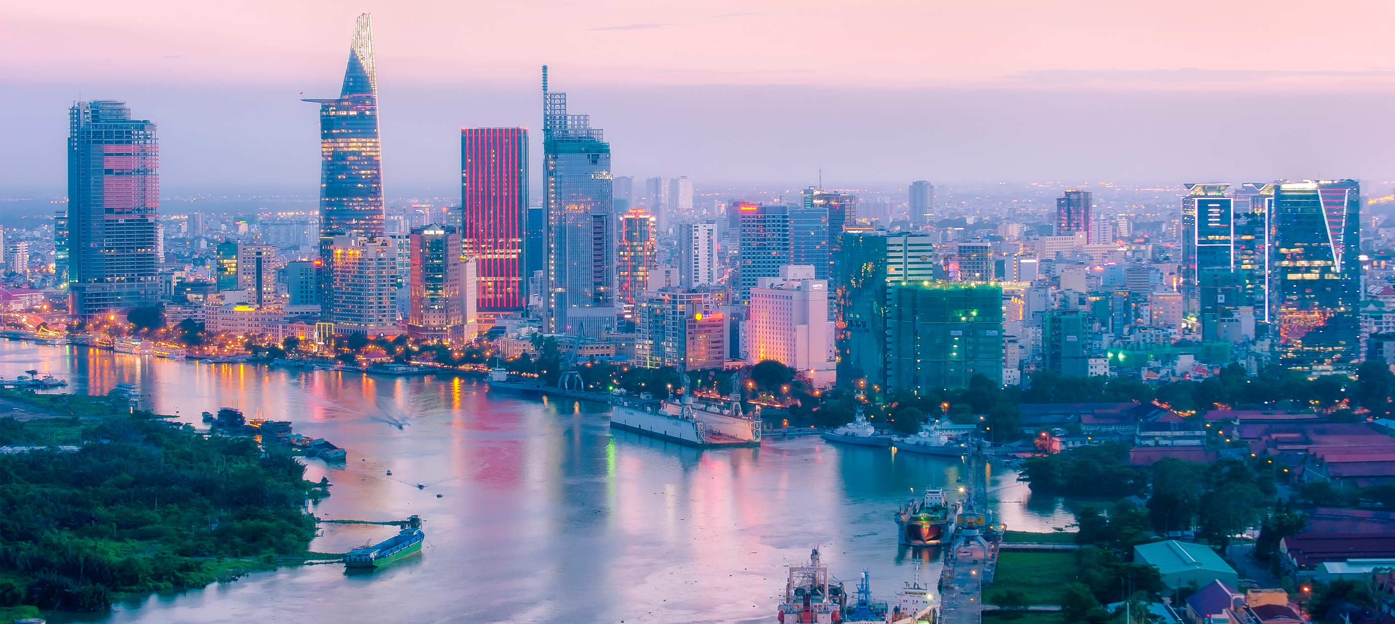 Ho Chi Minh City skyline at dusk with high-rise buildings along the Saigon River and boats moving through the water.