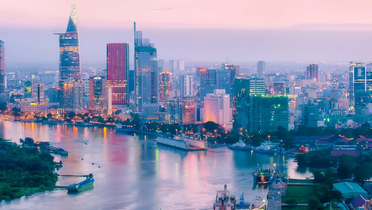 Ho Chi Minh City skyline at dusk with high-rise buildings along the Saigon River and boats moving through the water.