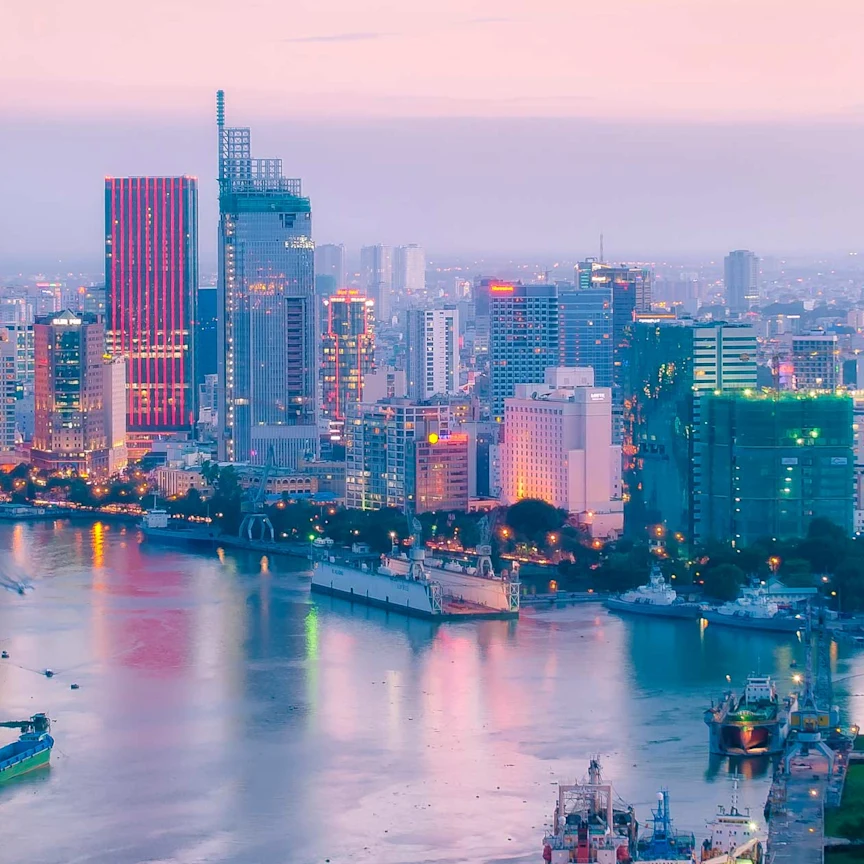 Ho Chi Minh City skyline at dusk with high-rise buildings along the Saigon River and boats moving through the water.