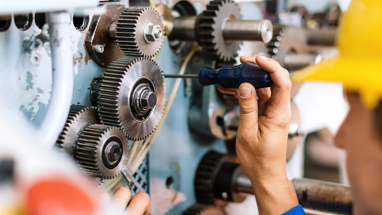 Industrial worker using a screwdriver to perform maintenance on gears and machinery in a manufacturing facility.