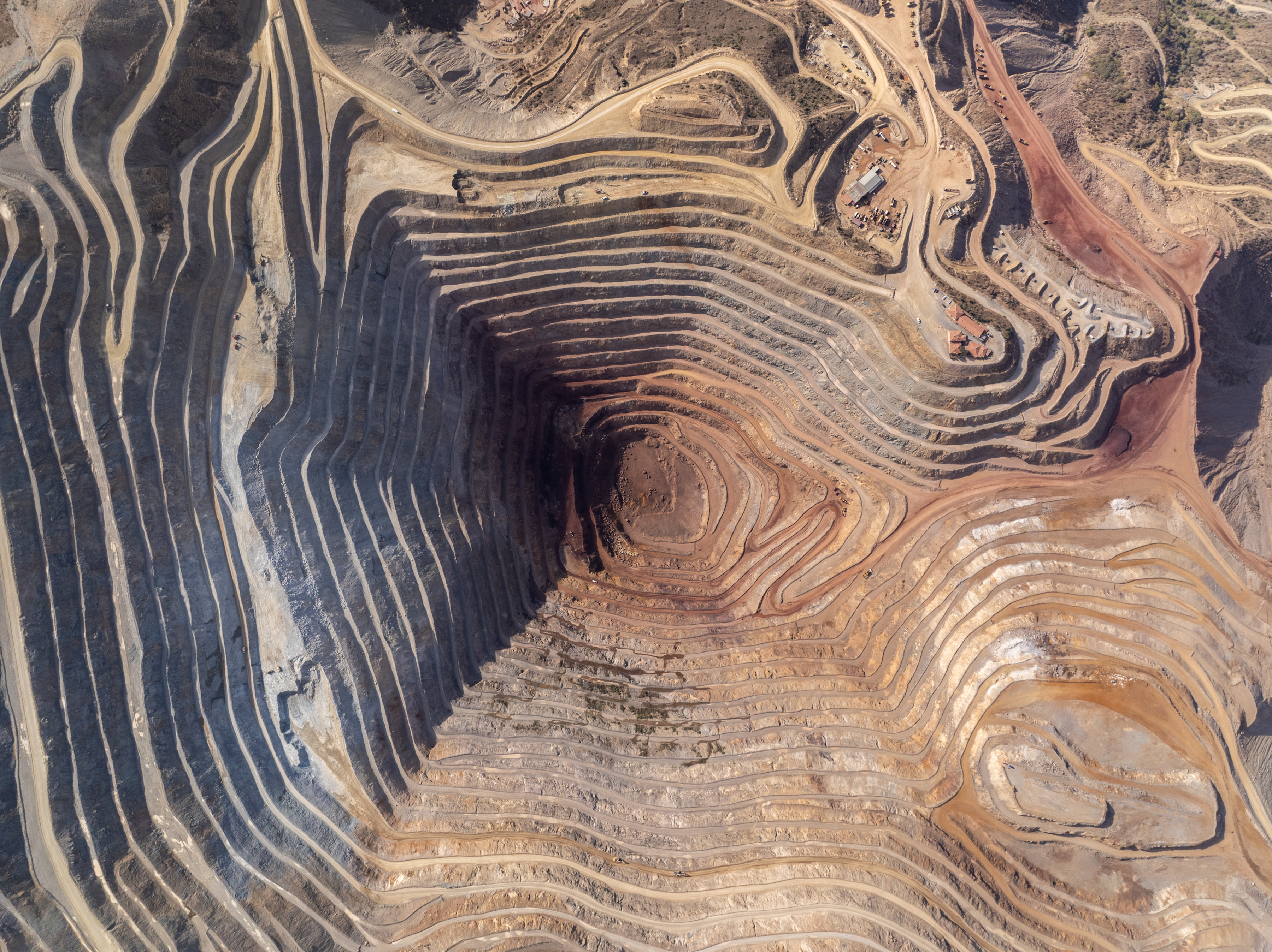 Aerial view of an open-pit mine with terraced layers carved into the earth, showing shades of brown and gray soil with winding access roads.