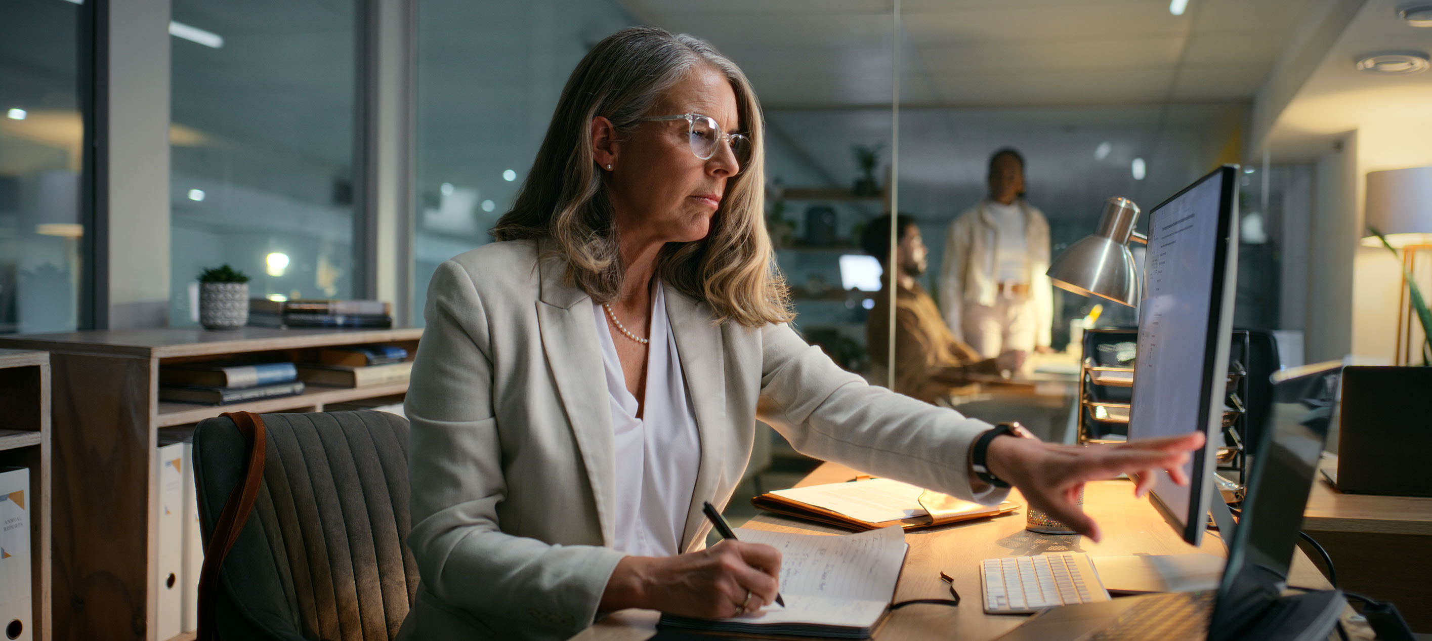 Businesswoman wearing glasses writing in a notebook while using a desktop computer at an office desk with colleagues in background.