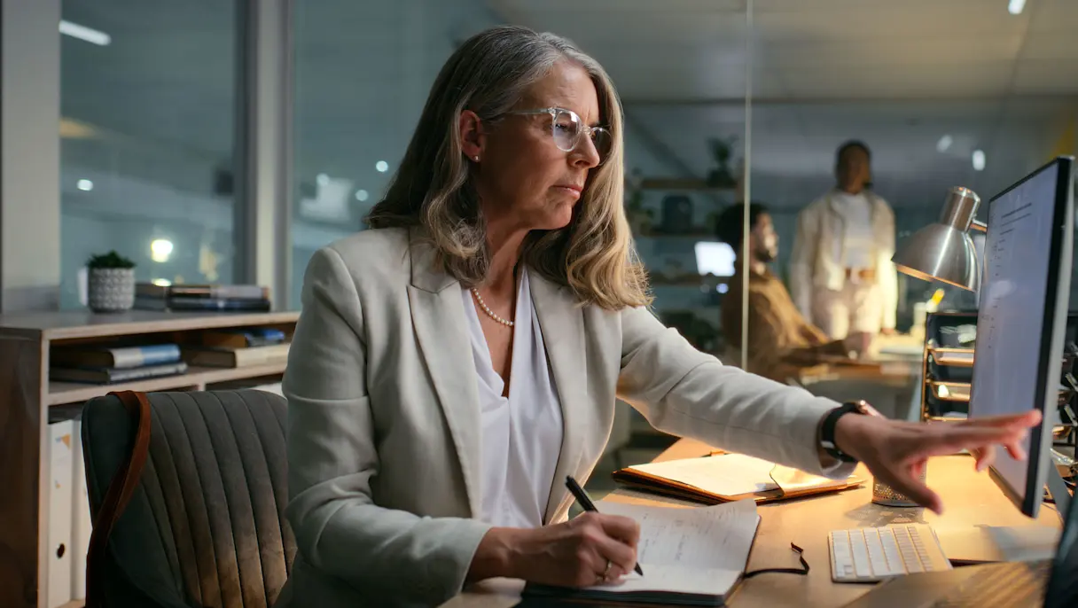 Businesswoman wearing glasses writing in a notebook while using a desktop computer at an office desk with colleagues in background.