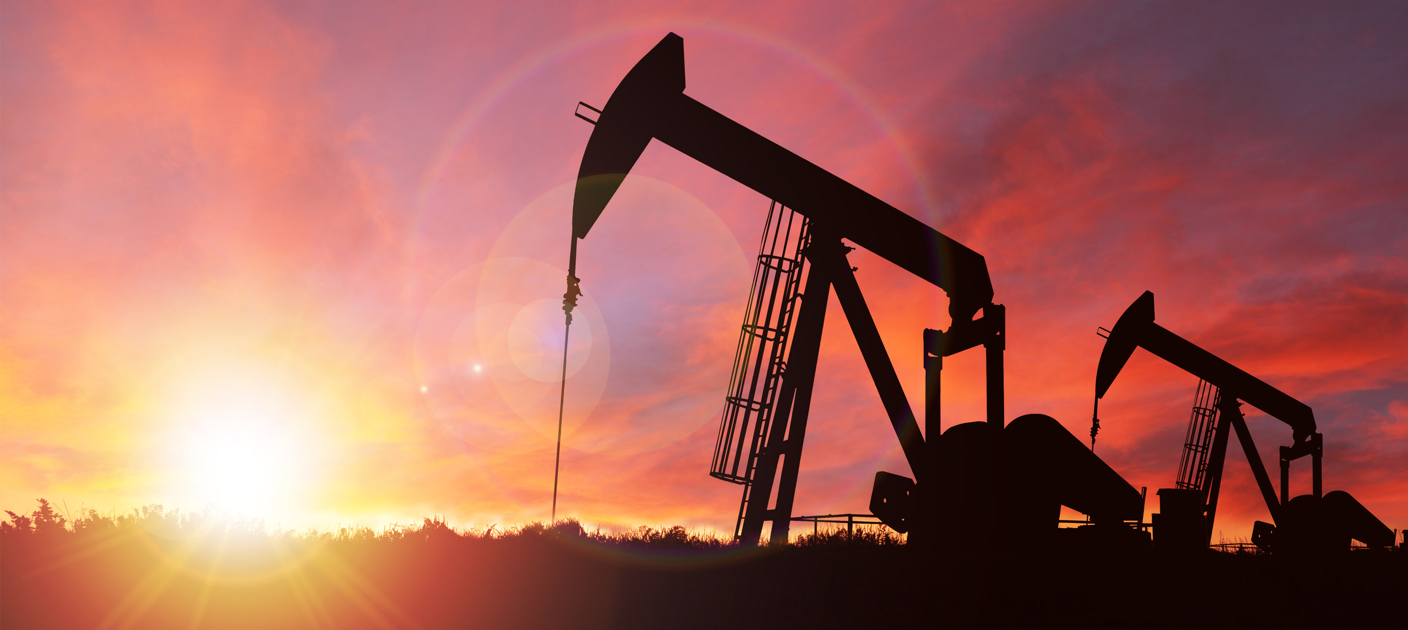 Silhouettes of two oil pump jacks operating against a vibrant orange and purple sunset sky with the sun near the horizon.