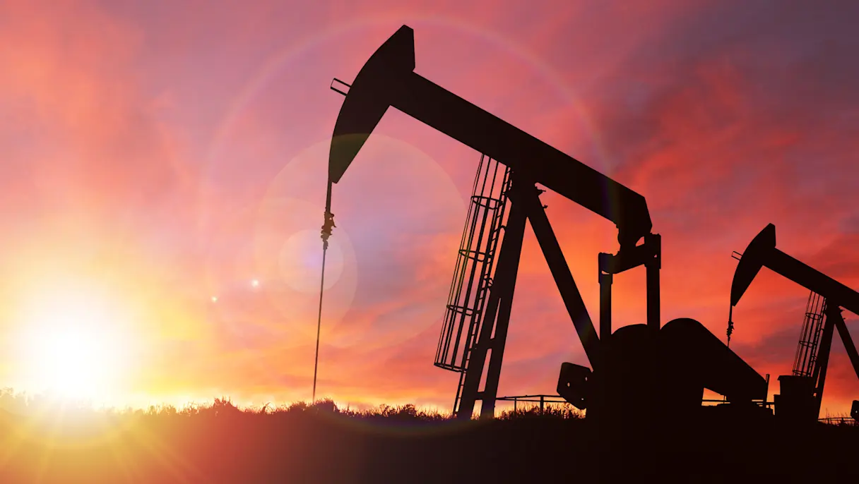 Silhouettes of two oil pump jacks operating against a vibrant orange and purple sunset sky with the sun near the horizon.