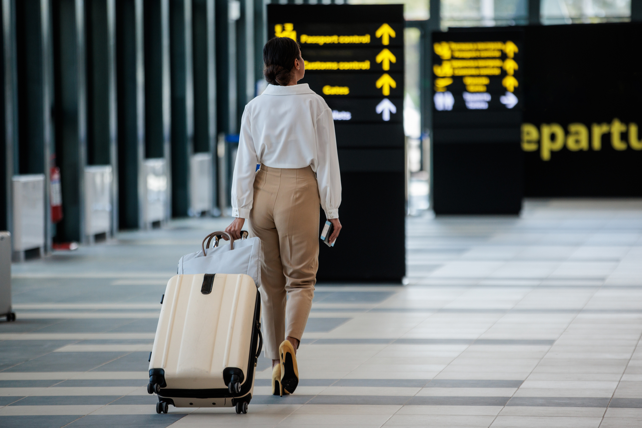 Woman in beige pants and white shirt walking in airport terminal, pulling a beige suitcase and holding a phone, with directional signs for passport and customs control ahead.