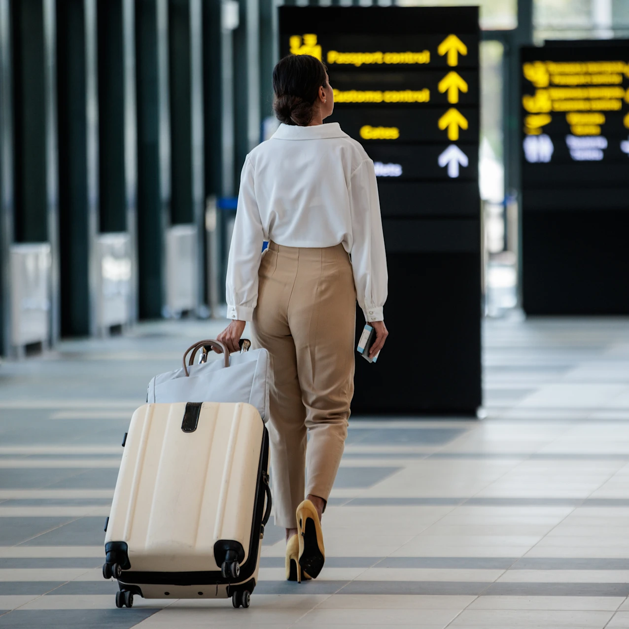 Woman in beige pants and white shirt walking in airport terminal, pulling a beige suitcase and holding a phone, with directional signs for passport and customs control ahead.
