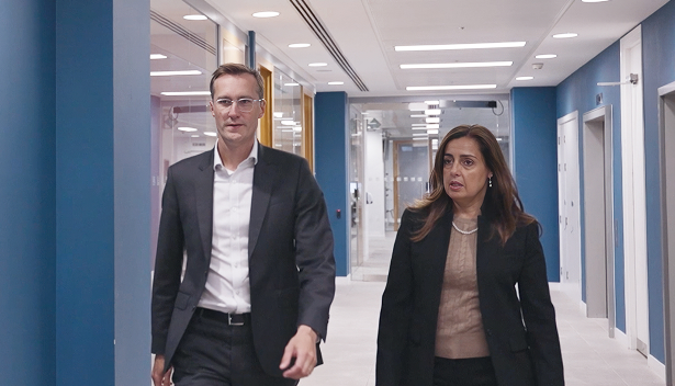 Two business professionals, a man and a woman, walking down a brightly lit office corridor with blue walls and white doors.