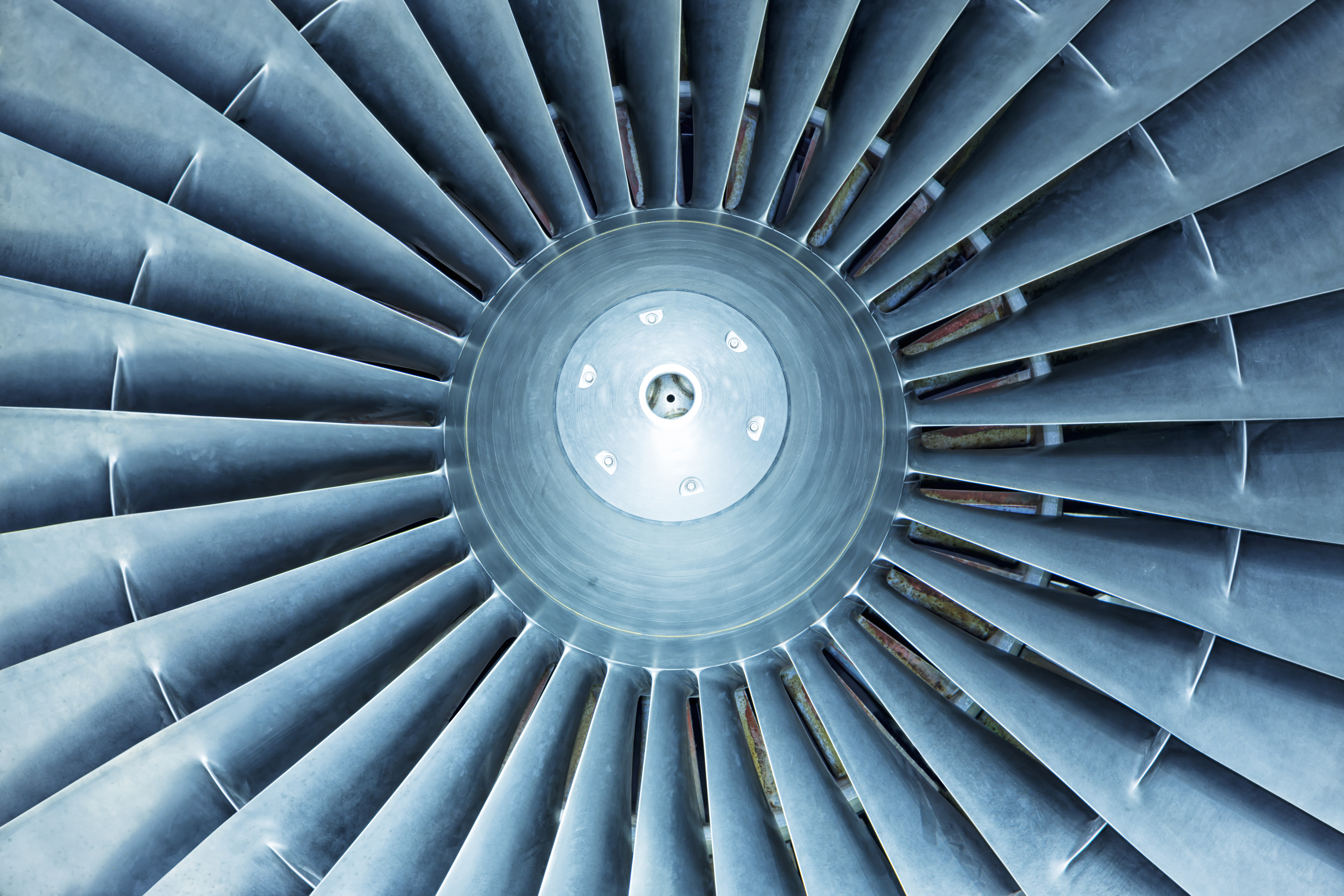 Close-up view of the metallic blades of a jet engine fan arranged in a circular pattern, showing details of the central hub and blades in blue-gray tones.