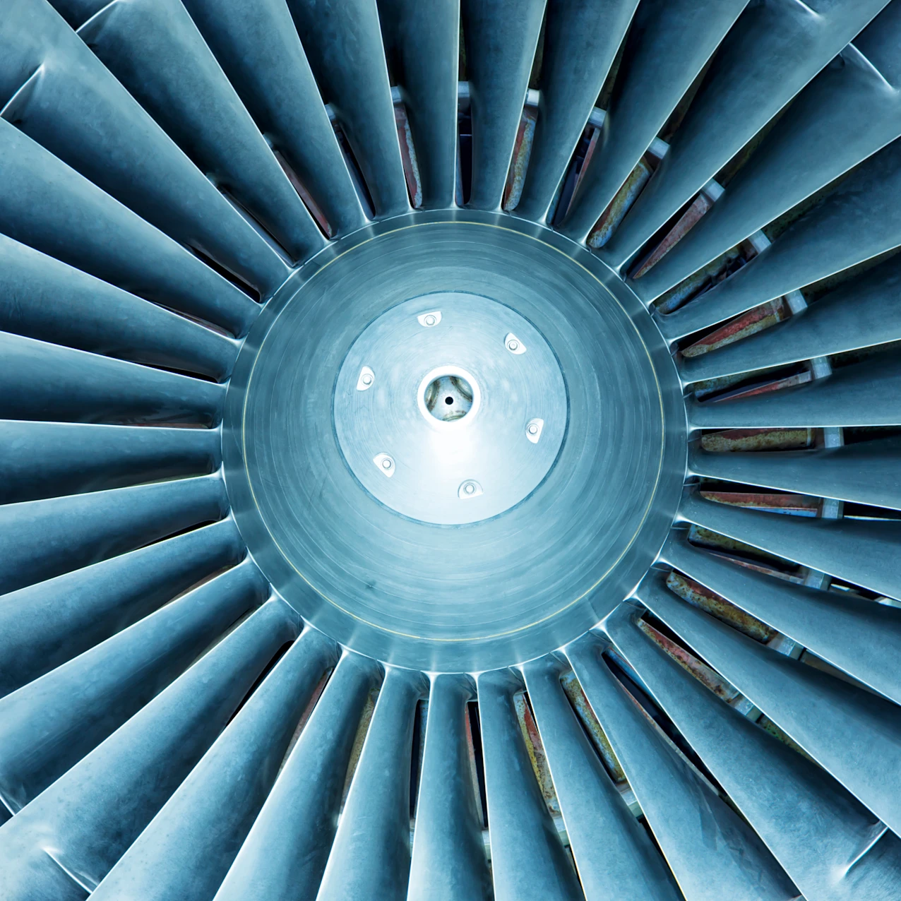 Close-up view of the metallic blades of a jet engine fan arranged in a circular pattern, showing details of the central hub and blades in blue-gray tones.