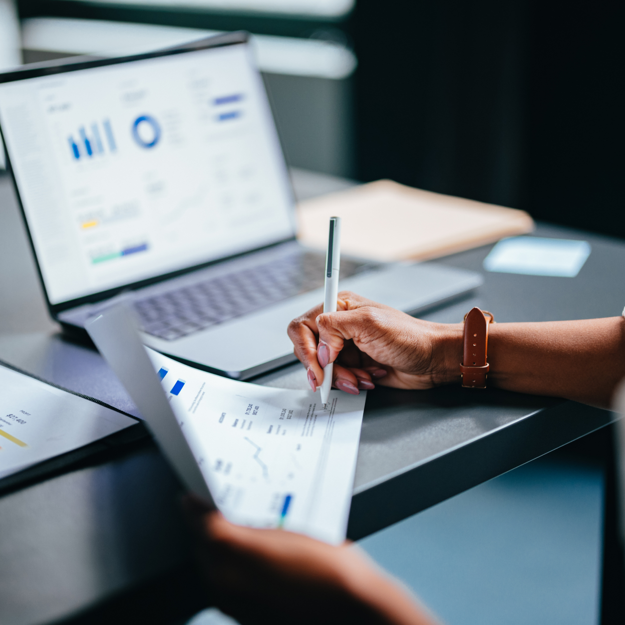 Person reviewing and marking financial documents with charts and graphs beside an open laptop displaying data on a desk.