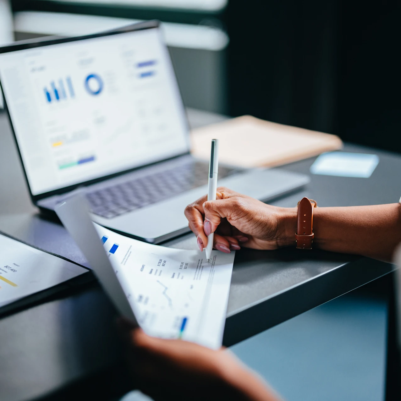 Person reviewing and marking financial documents with charts and graphs beside an open laptop displaying data on a desk.