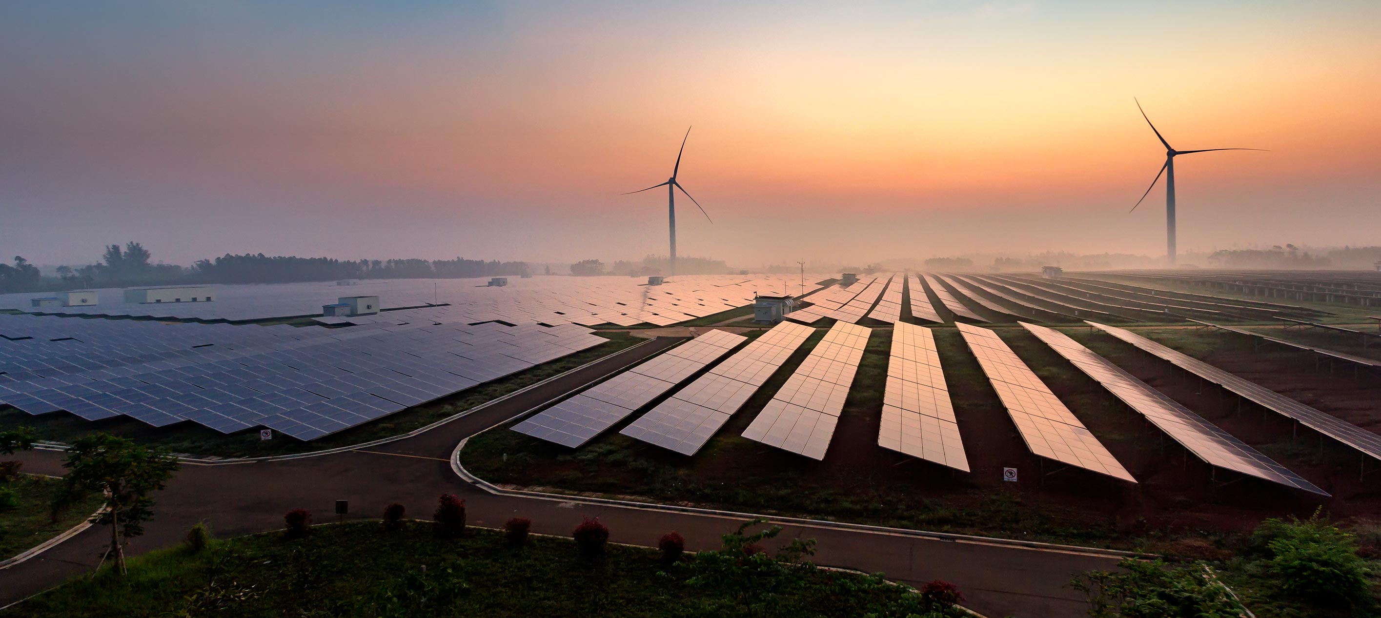 Expansive solar farm with rows of photovoltaic panels and two wind turbines against a misty sunrise, symbolising renewable energy. 