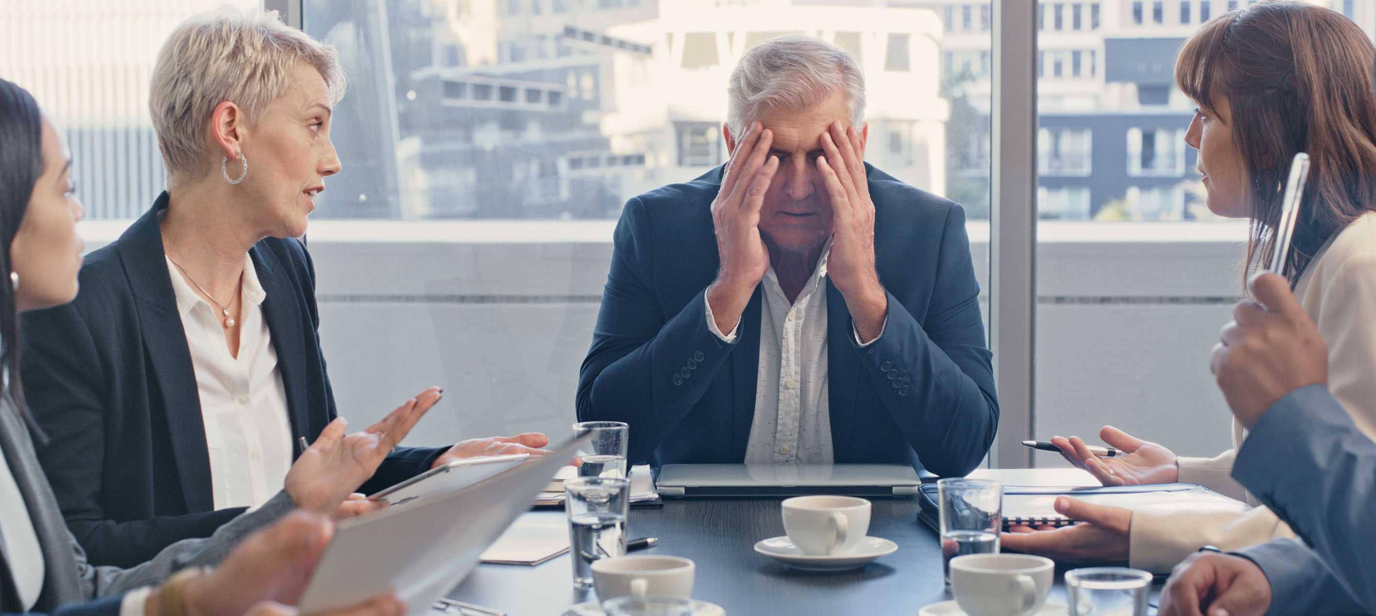 Business professionals in a tense boardroom meeting with an older male executive holding his head in his hands.