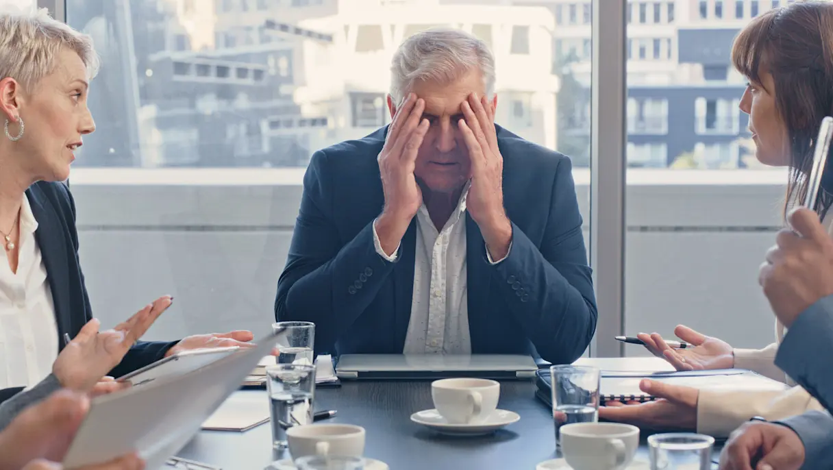 Business professionals in a tense boardroom meeting with an older male executive holding his head in his hands.