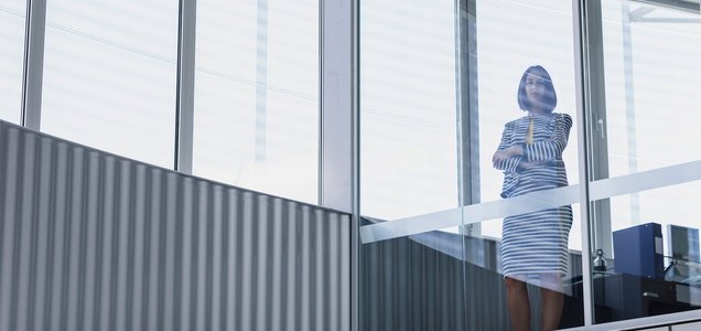 A woman in a striped dress stands with arms crossed beside floor-to-ceiling windows in a modern office space.