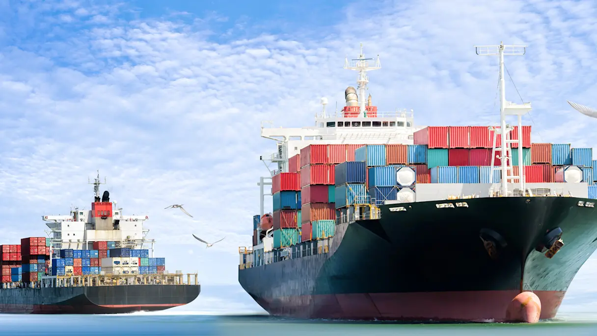 Two cargo ships carrying colorful shipping containers sail across the sea under a blue, cloudy sky, illustrating global trade and maritime transport.