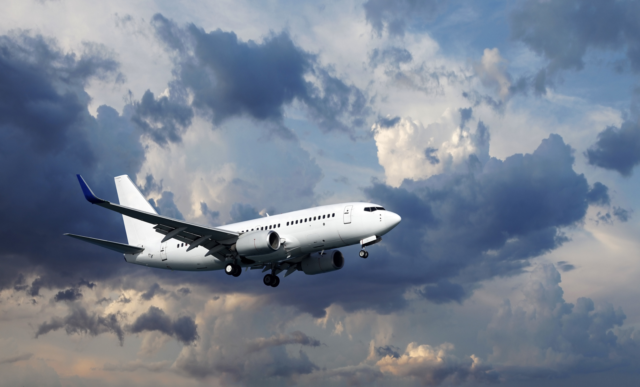 White passenger airplane flying with landing gear down against a dramatic cloudy sky at dusk.