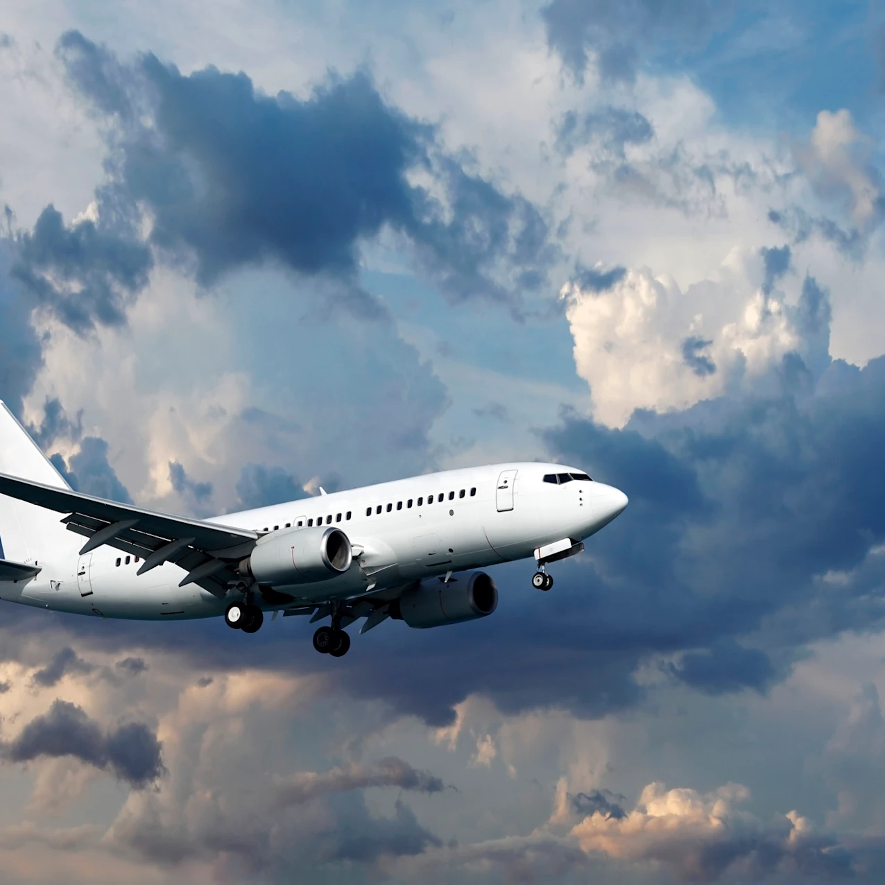 White passenger airplane flying with landing gear down against a dramatic cloudy sky at dusk.