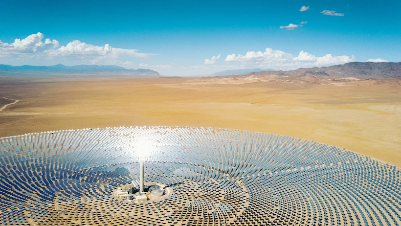 Aerial view of a solar power tower surrounded by heliostats in a desert landscape under a clear blue sky with distant mountains.