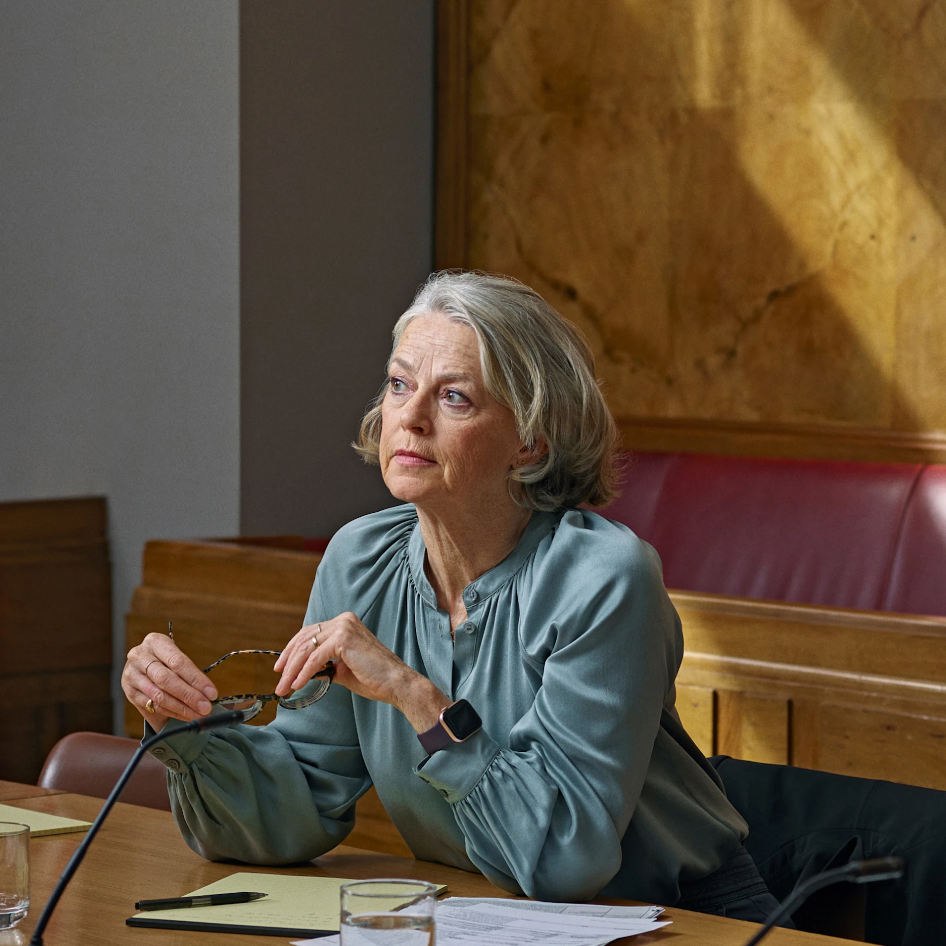 Person in teal blouse seated at conference table with papers and microphone, holding glasses in a wood-paneled room.