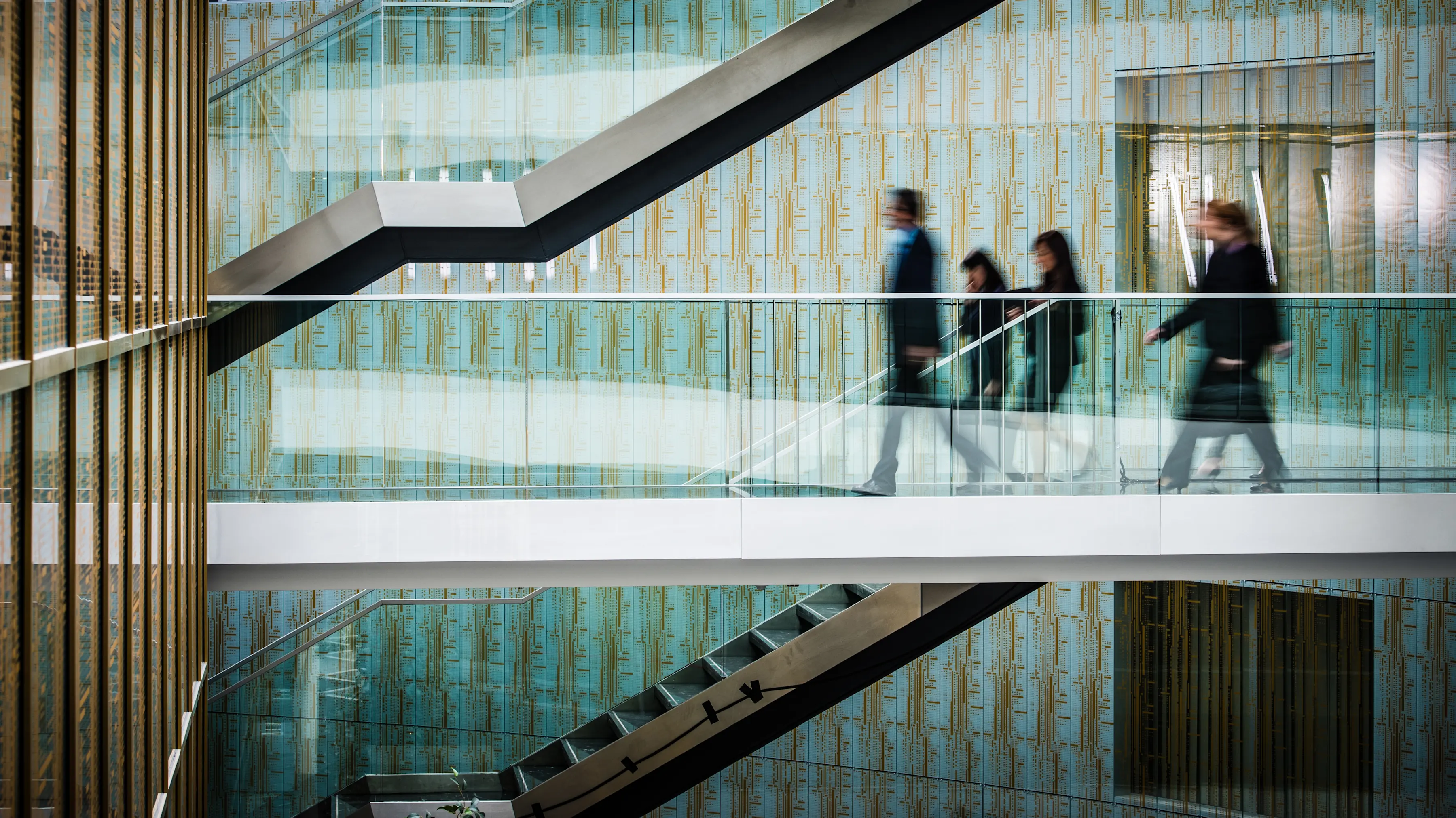 Blurred motion of four people walking on a glass-walled corridor inside a modern building with patterned blue and gold walls and a black staircase.