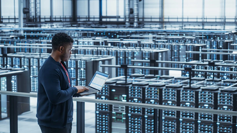 A man operates a laptop in a spacious server room filled with servers and networking equipment.