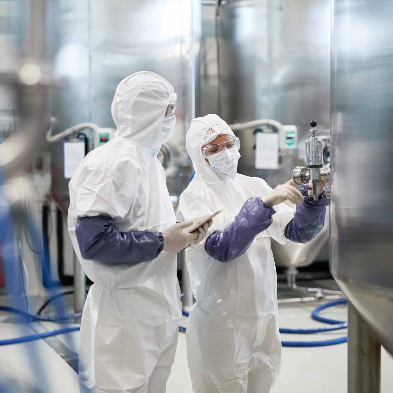 Two workers in full white protective suits and gloves operate a large stainless steel tank in a clean industrial facility.