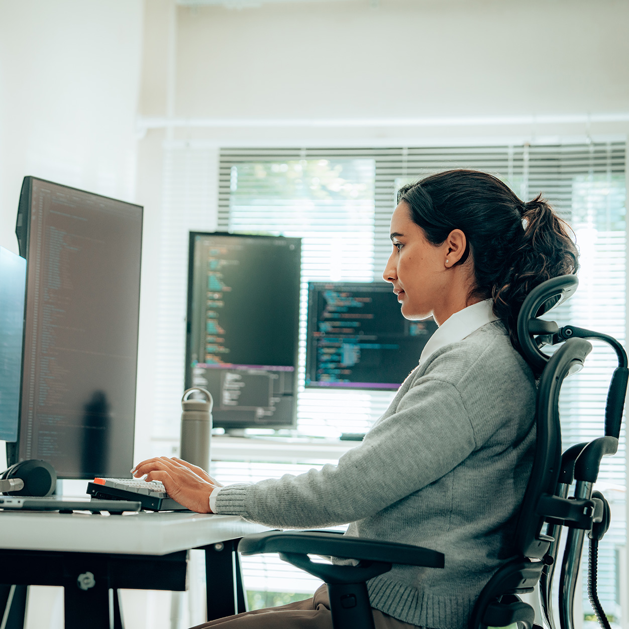 Person in gray sweater working on computer coding at desk with multiple monitors and office chair in bright modern room.