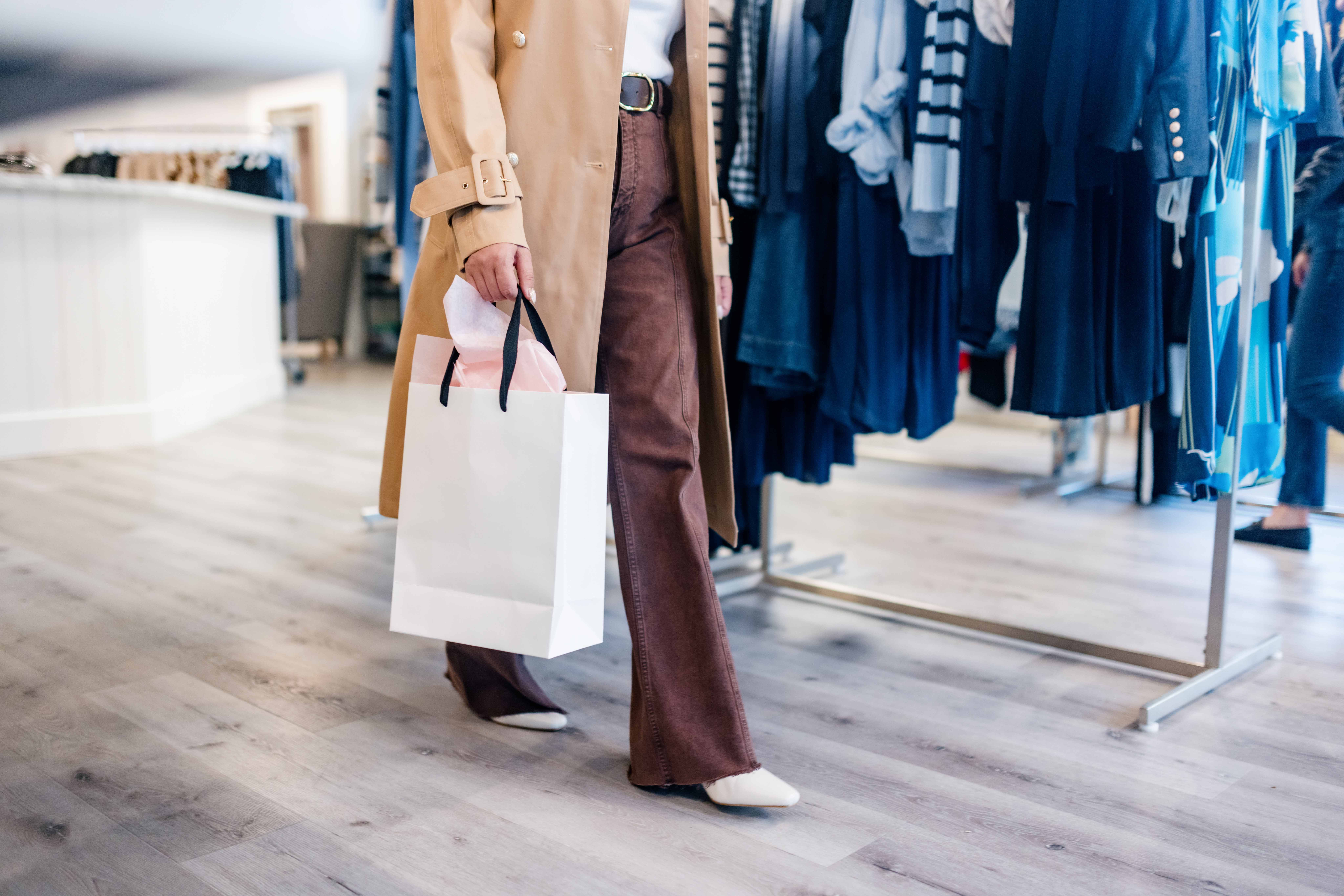 Person wearing brown pants and a beige coat holding a white shopping bag while walking inside a clothing store with racks of clothes.