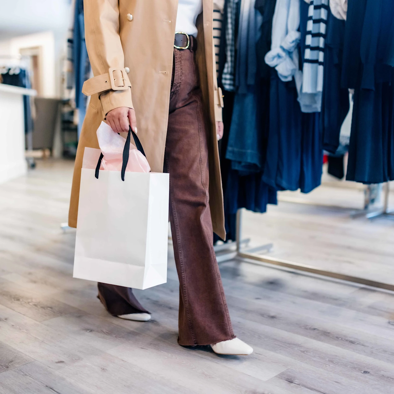 Person wearing brown pants and a beige coat holding a white shopping bag while walking inside a clothing store with racks of clothes.