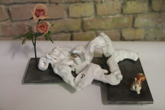 A man form in Fayence lying on two plates of aluminium, a plastic flower is over his head, and a kitsch porcelain cat is playing at his feet