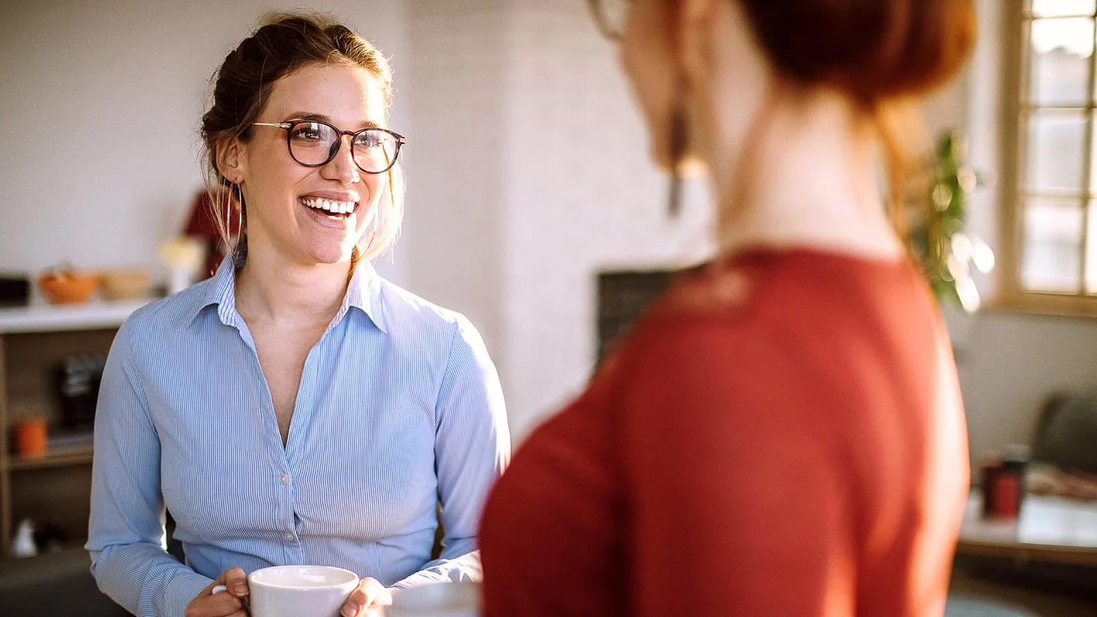 Person wearing glasses and a light blue button-down shirt holding a white mug while speaking with another person in a bright indoor setting.