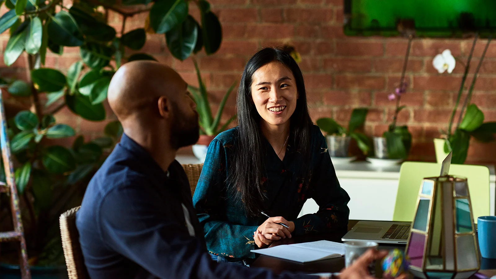 Two coworkers seated at a table discussing work beside a laptop and notebook in a plant-filled office with exposed brick walls.