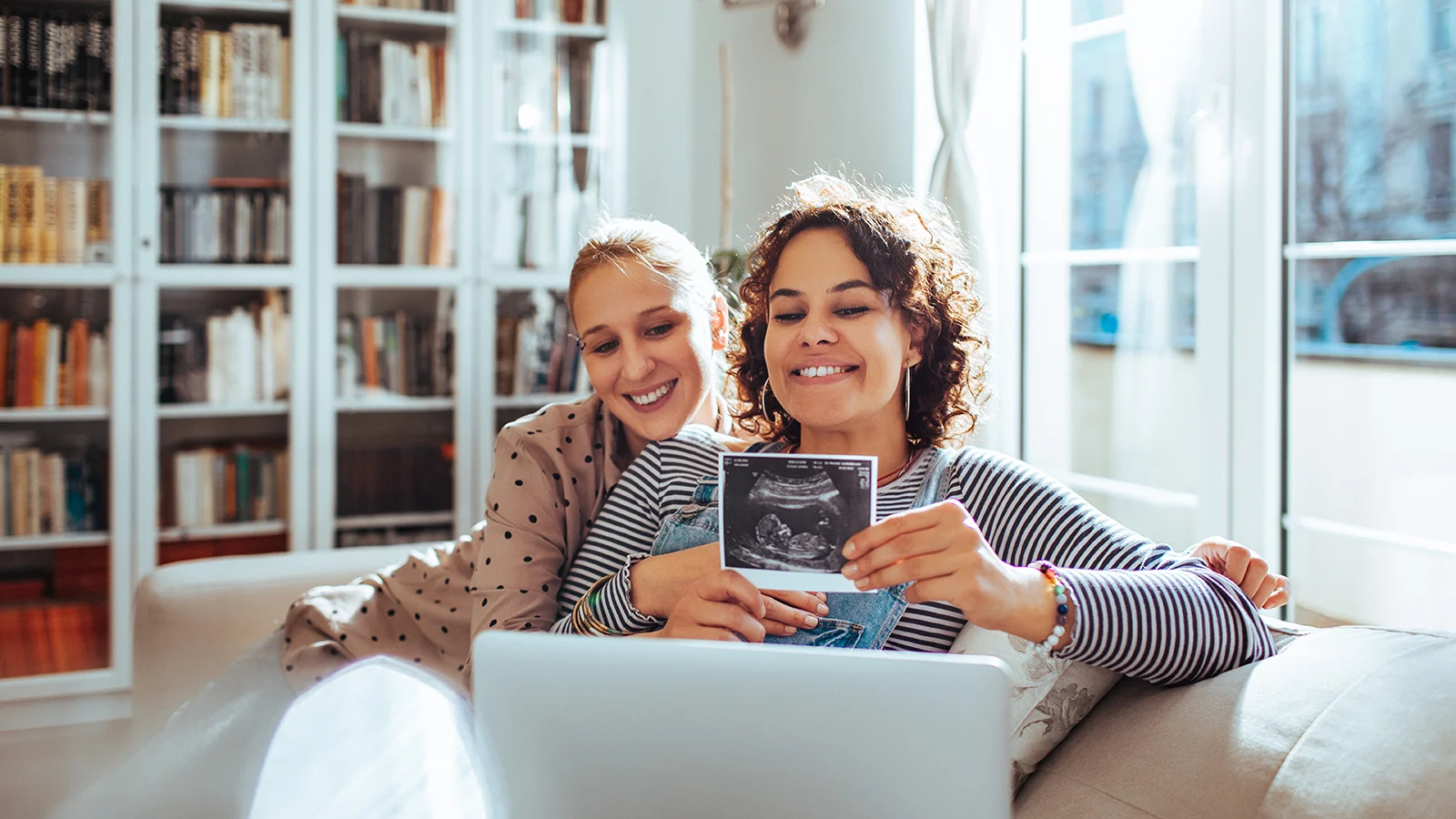 Two people sit closely together on a sofa in a bright room, holding an ultrasound image in front of an open laptop. Bookshelves and large sunlit windows fill the background.