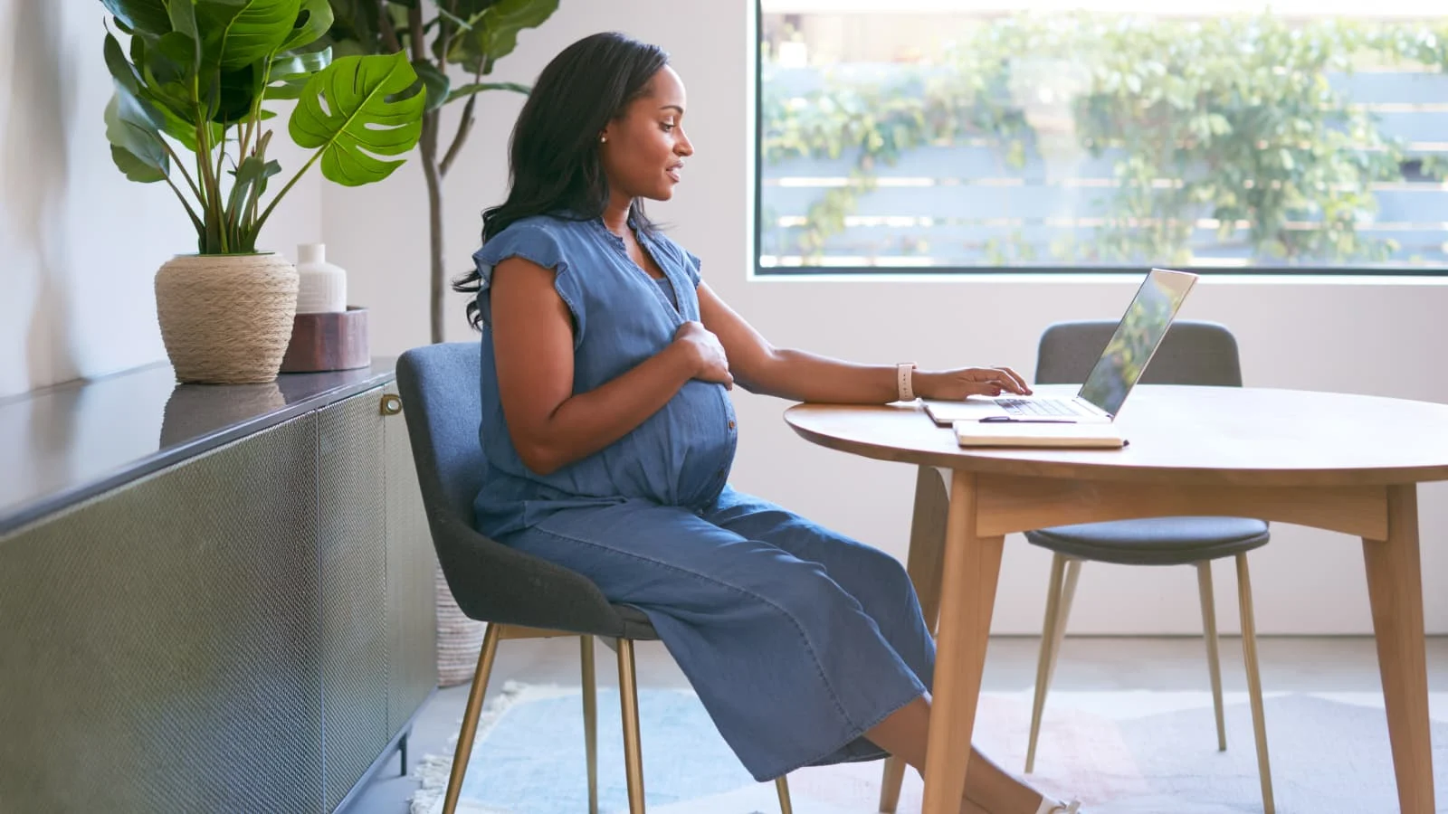 Pregnant woman seated at a dining table using a laptop in a bright home workspace, with a notebook on the table and large indoor plants nearby.