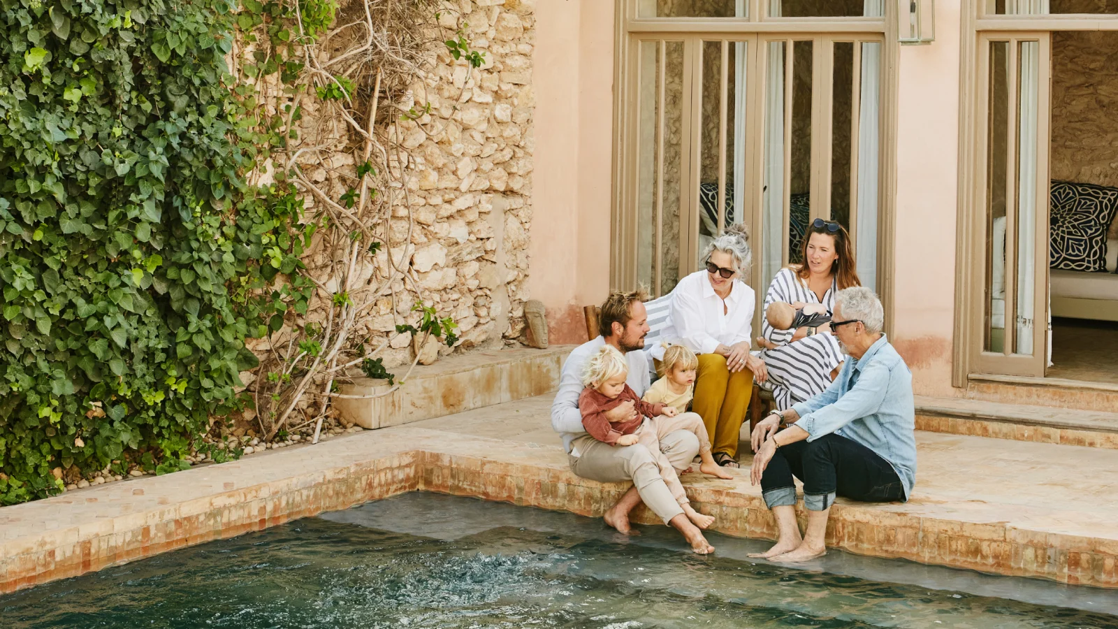 Next-Gen Wealth - Overview - Image 1 Group of adults and children sitting beside a courtyard pool outside a stone house.