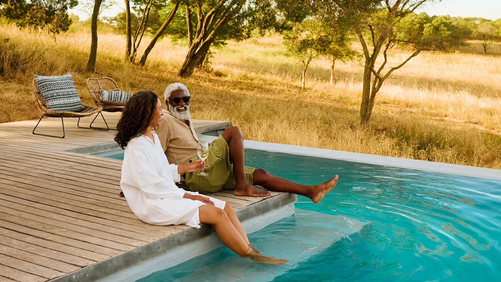 Two people sit on the edge of an outdoor pool with their feet in the water, surrounded by a wooden deck, trees, and a natural landscape.