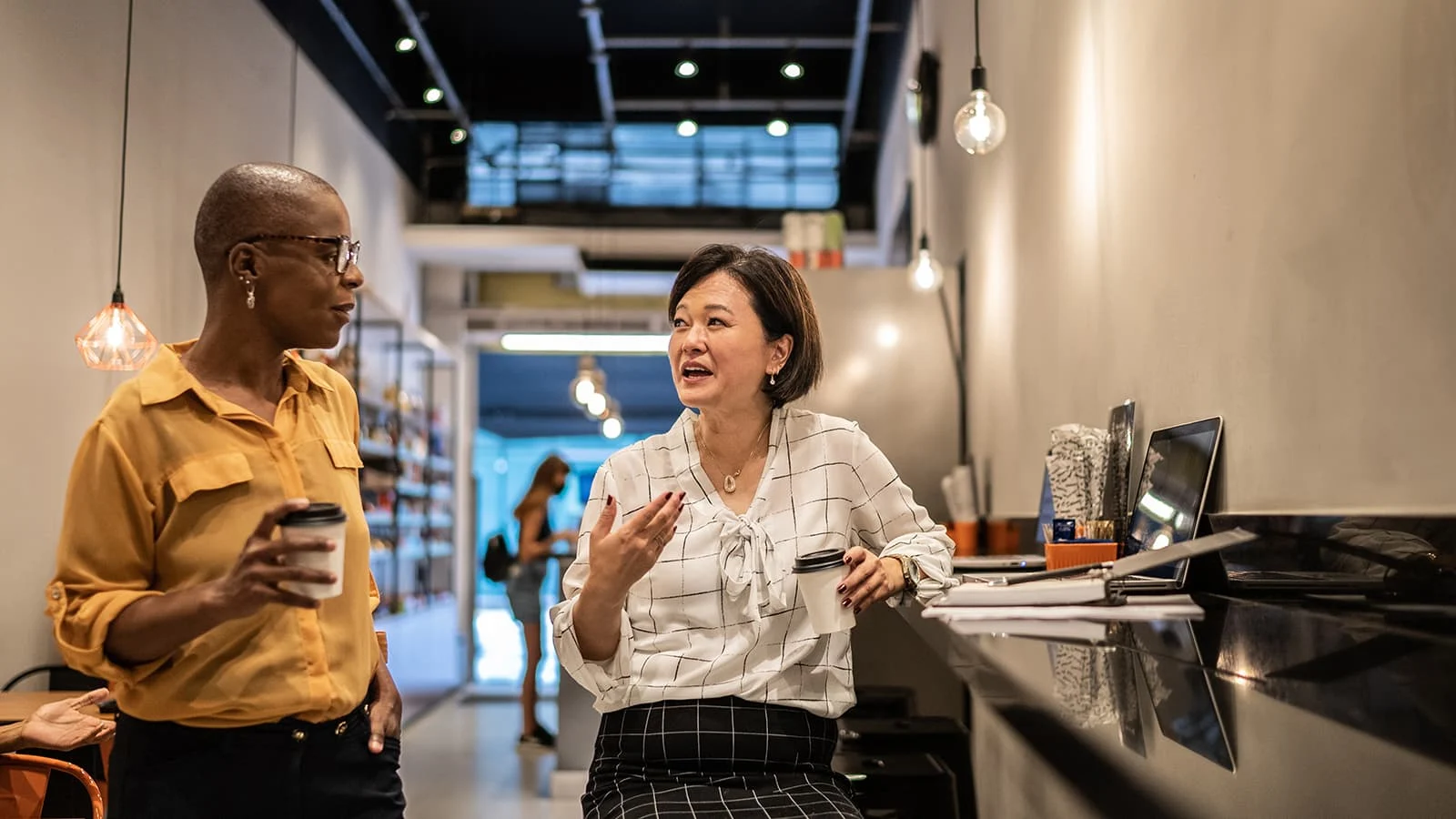Two coworkers chatting over coffee in a modern office café, with an open laptop on a counter nearby.