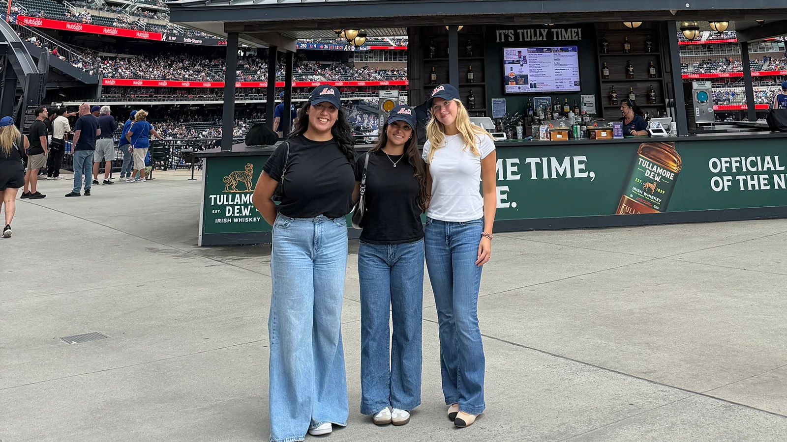 Three people pose together at a baseball stadium in front of a concourse bar, wearing matching baseball caps.