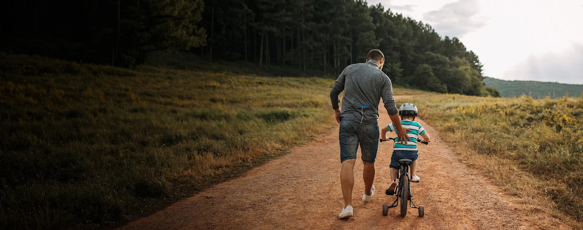 A father helps his son ride a bike down a path in a field.